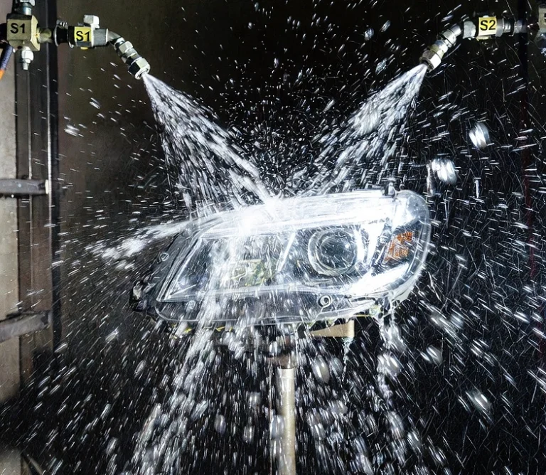 Automotive headlight being tested inside the rain spray test chamber with high-pressure water spray from S1/S2 nozzles hitting the part, realistic water droplets flying, dramatic lab lighting, professional testing scene photography.