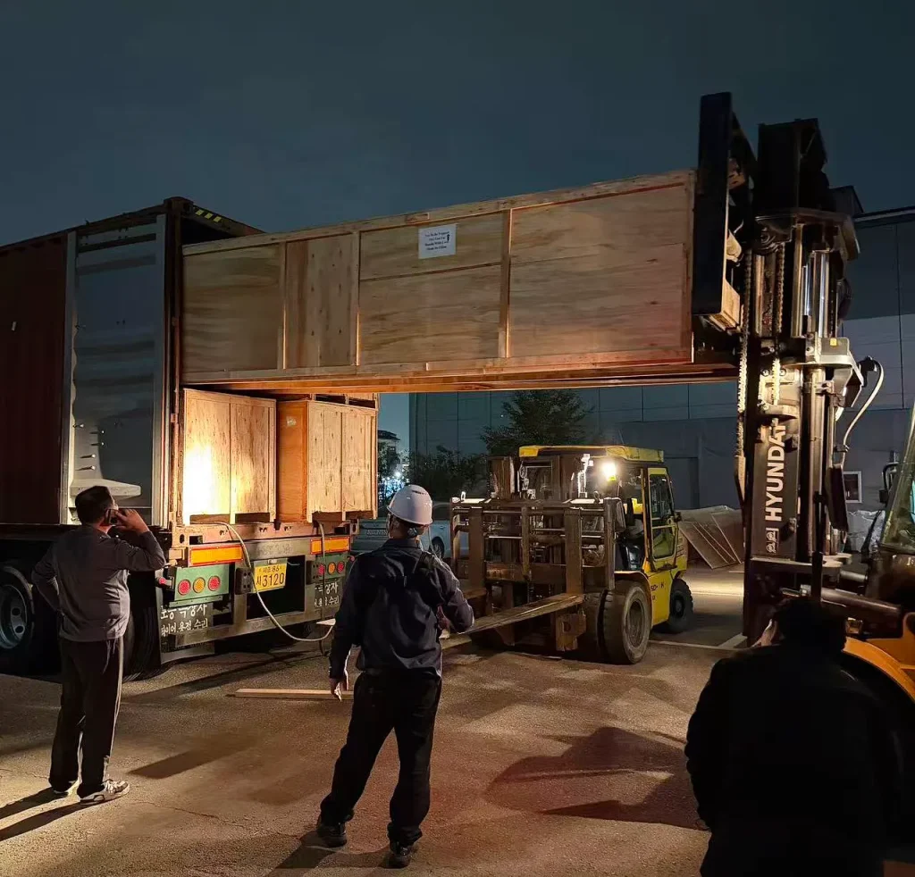 Heavy-duty forklift handling an oversized wooden shipping crate during nighttime unloading of KINGPO dust test chamber components in South Korea.