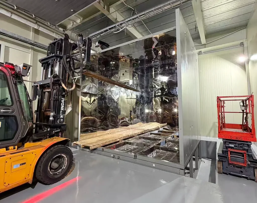 Forklift assisting with positioning a large structural section inside the stainless steel walk-in sand and dust test chamber during assembly at SGS Korea.